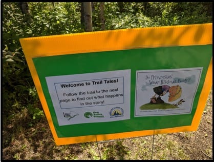 An orange sign with green posterboard in the middle. Pasted to the board are two sheets of white paper: the one on the left says "Welcome to Trail Tales! Follow the trail to the next page to find out what happens in the story." The page on the right is a picture of the cover of Do Princesses Wear Hiking Boots by Carmela Lavigna Coyle.