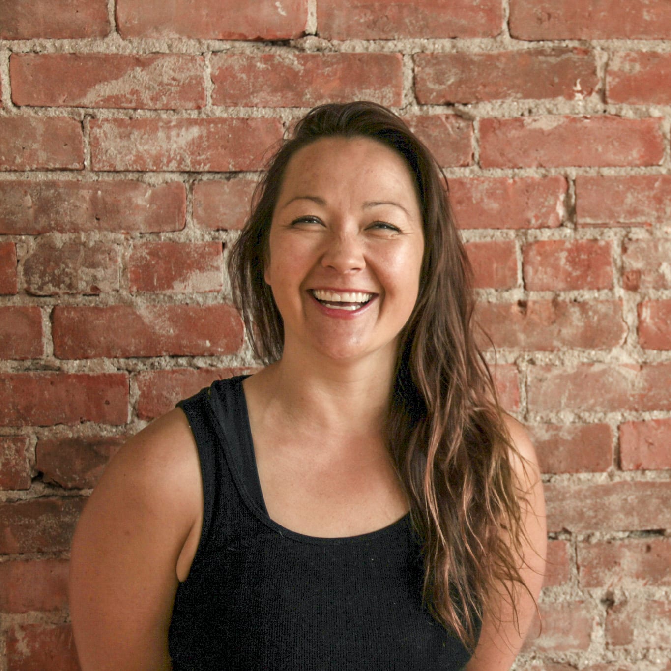 Shonna Lamb, wearing a black tank top, with long wavy brown hair, smiles while standing against a red brick wall.