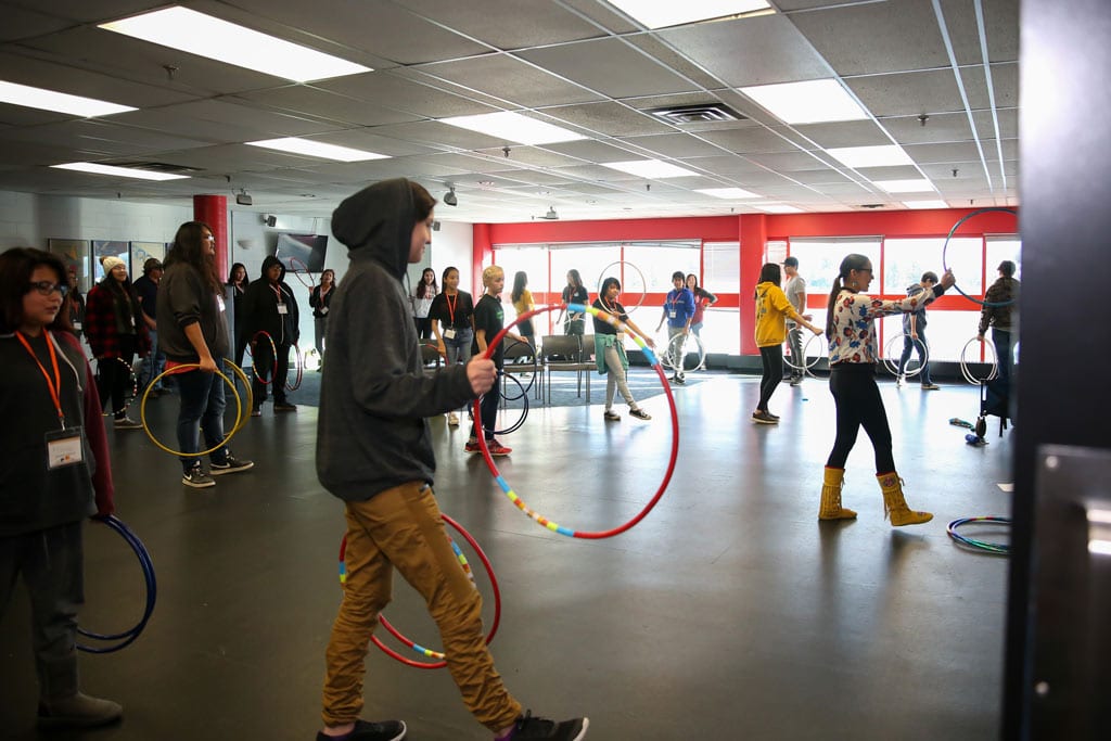 Sandra Lamouche teaches a hoop dance class to a group of youth. Sandra is seen on the right side of the image holding a hoop at eye level while the youth are seen filling the room copying her.