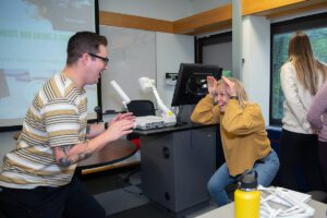 Two people are playing Giants, Wizards, Elves – a game to help teach the Phys Ed and Wellness section of the Alberta K-6 Curriculum. The person on the left is posed like a wizard with fingers pointed like magic wands. The person on the right is crouched down in an elf pose using hands for elf ears.