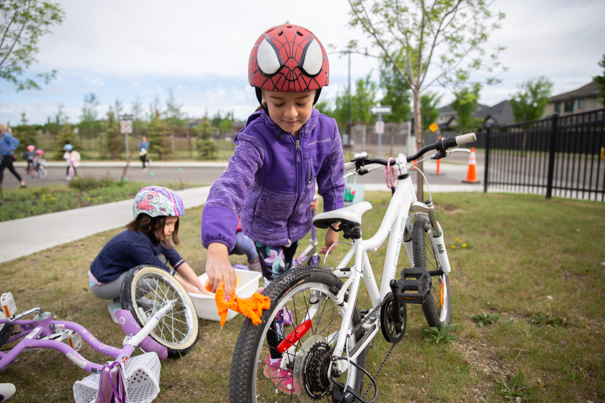 A young girl wearing a purple sweater and a Spiderman cycling helmet washes her white bike.